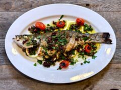 Life At The Table Grilled Branzino with Mediterranean Gremolata. A whole grilled branzino on a white plate garnished with kalamata olives, capers, lemon juice olive oil, and blistered grape tomatoes on a white plate sitting on a wooden table.