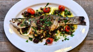 Life At The Table Grilled Branzino with Mediterranean Gremolata. A whole grilled branzino on a white plate garnished with kalamata olives, capers, lemon juice olive oil, and blistered grape tomatoes on a white plate sitting on a wooden table.