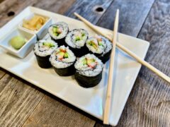 Life At The Table California Roll | A California Roll on a white plate cut into 6 pieces with a ball of wasabi and pickled ginger on the plate along with chopsticks.