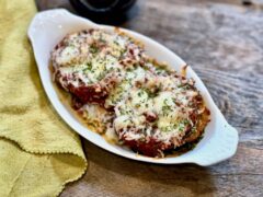 Life At The Table Eggplant Parmigiana. Eggplant parmigiana in a white dish sitting on a wooden table with a green napkin and a glass of wine in the background.