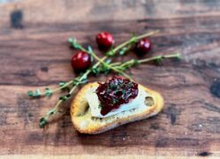 Life At The Table Shrimp and Cranberry Crostini. A shrimp and cranberry crostini on a wooden serving tray surrounded by fresh thyme and cranberries.