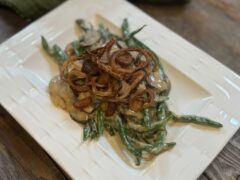 Life At The Table Green Bean Casserole. Green bean casserole with fresh green beans on a white plate garnished with fried shallots on a wooden table.