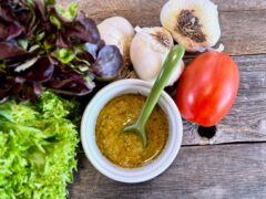 Life At The Table Garlic Vinaigrette. Garlic vinaigrette in a white bowl with a green spoon on a wooden table surrounded by lettuce, heads of garlic, and a Roma tomato.