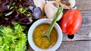 Life At The Table Garlic Vinaigrette. Garlic vinaigrette in a white bowl with a green spoon on a wooden table surrounded by lettuce, heads of garlic, and a Roma tomato.