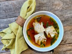 Life At The Table Wonton Soup. A bowl of wonton soup with three wontons and garlnished with chopped green onions sitting on a wooden table with a green napkin beside it.