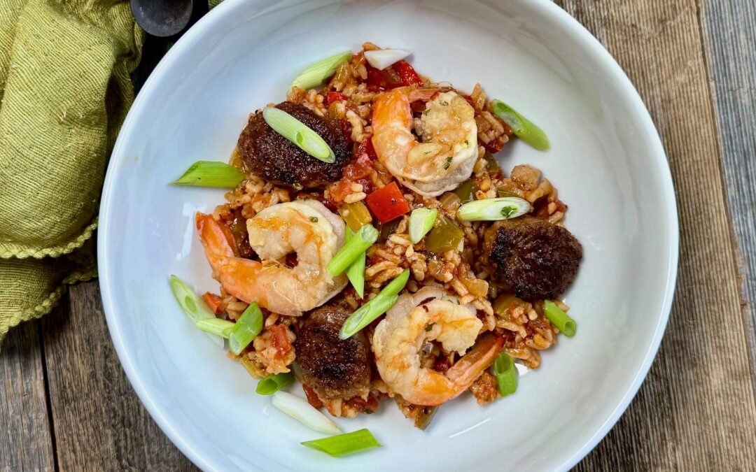 Life At The Table Jambalaya. A white bowl of jambalaya with sausage, shrimp and chicken garnished with green onions with a green napkin next to the bowl on a wooden table.