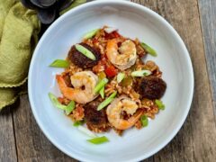 Life At The Table Jambalaya. A white bowl of jambalaya with sausage, shrimp and chicken garnished with green onions with a green napkin next to the bowl on a wooden table.