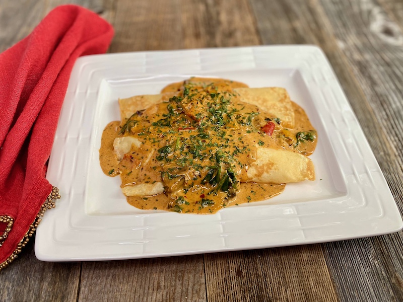 Life At The Table Savory and Sweet Crepes Virtual Cooking Class. Two crepes with Cajun sauce on a rectangular white plate sitting on a wooden table with a red napkin beside it.