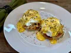 Life At The Table Crab Eggs Benedict. Crab Eggs Benedict on a white plate sitting on a wooden table with a green napkin beside it.