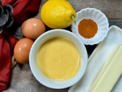 Life At The Table Hollandaise Sauce. A small bowl of hollandaise sauce surrounded by two eggs, one lemon, one stick of butter and cayenne with a red napkin just barely visible to the left, sitting on a wooden table.