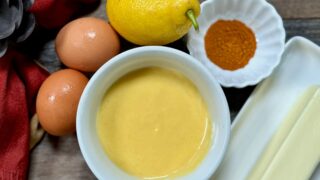 Life At The Table Hollandaise Sauce. A small bowl of hollandaise sauce surrounded by two eggs, one lemon, one stick of butter and cayenne with a red napkin just barely visible to the left, sitting on a wooden table.