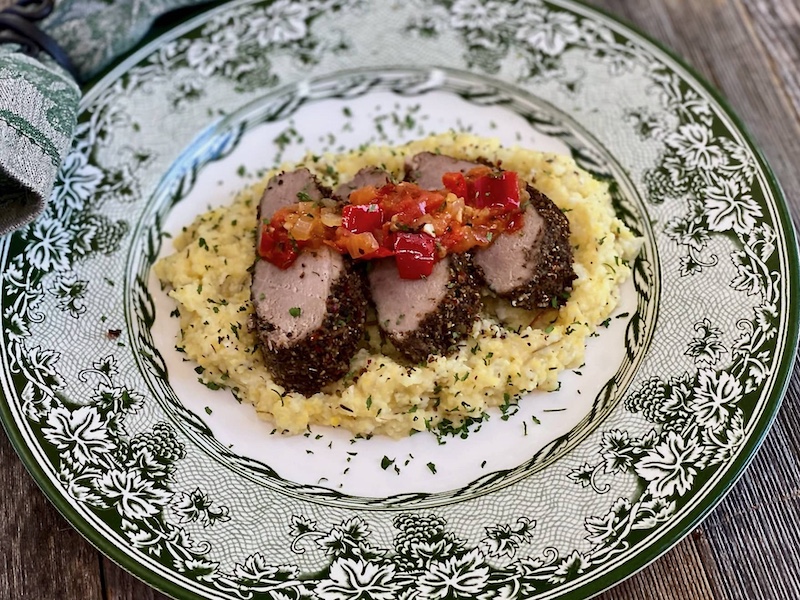 Life At The Table Jolly Juniper Roasted Pork Tenderloin Virtual Cooking Class. Three sliced pieces of Jolly Juniper Roasted Pork Tenderloin sitting on a bed of polenta on a green plate with a matching napkin beside it.