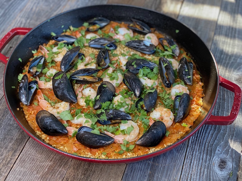 Life At The Table Paella Virtual Cooking Class. A pan of paella sitting on a wooden table. The paella is topped with shrimp and mussels.
