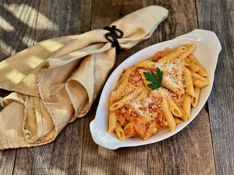 Life At The table Pasta alla Penne Virtual Cooking Class. A dish of pasta alla Penne on a wooden table with a yellow napkin lying beside it.