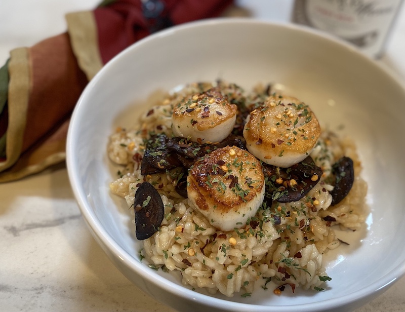 Life At The Table Risotto Virtual Cooking Class. A bowl of risotto with scallops and mushrooms on a wooden table with a napkin beside it.
