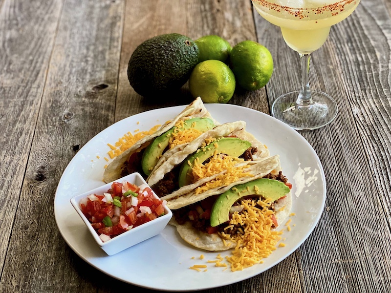 Life At The Table Virtual Taco Cooking Class. A plate of three tacos with salsa fresca on a white plate with a margarita sitting next to it on a wooden table.
