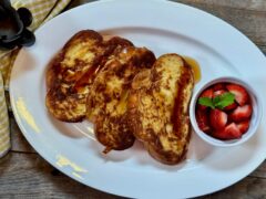 Life At The Table French Toast. A white with three slices of french toast made from challah bread with a small bowl of strawberries garnished with a mint sprig sitting on a wooden table with a yellow napkin.