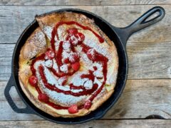Life At The Table Dutch Baby. A Dutch baby in a cast iron skillet sitting on a wooden table garnished with raspberry coulis, raspberries, and powdered sugar.