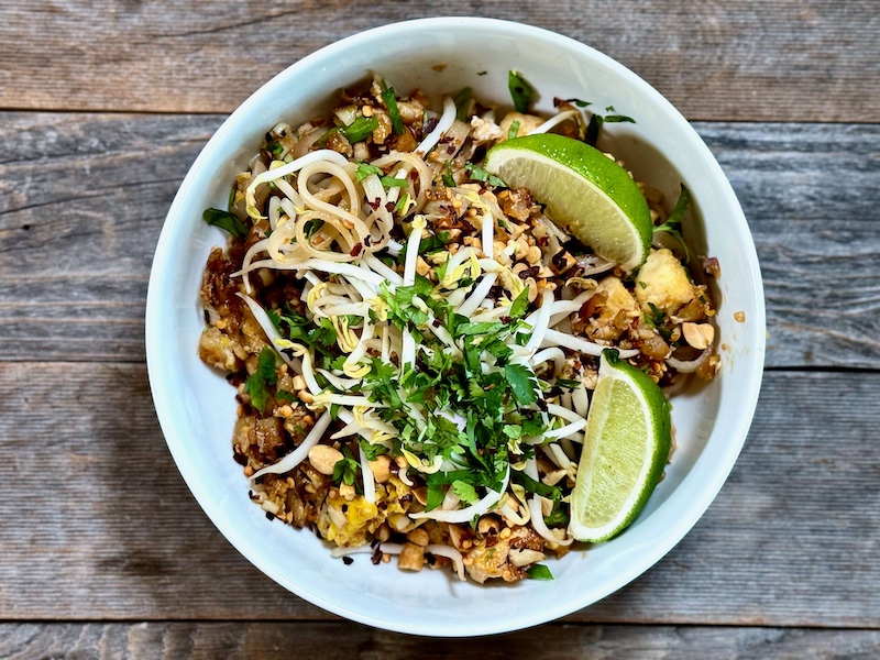 Life At The Table Pad Thai Culinary Team Building. A white bowl filled with Pad Thai and garnished with cilantro, peanuts, and lime wedges sitting on a wooden table.
