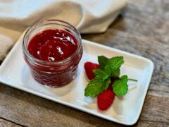 Life At The Table Raspberry Coulis. Raspberry coulis in a small glass canning jar sitting on a white plate sitting on a wooden table with fresh raspberries and a sprig of mint next to it. The white plate has a cream colored napkin next to it.