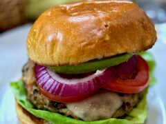 Life At The Table Chicken Burger. A chicken burger on a white plate topped with lettuce, red onion, tomato and avocado.