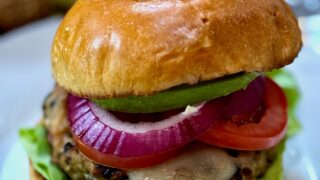 Life At The Table Chicken Burger. A chicken burger on a white plate topped with lettuce, red onion, tomato and avocado.