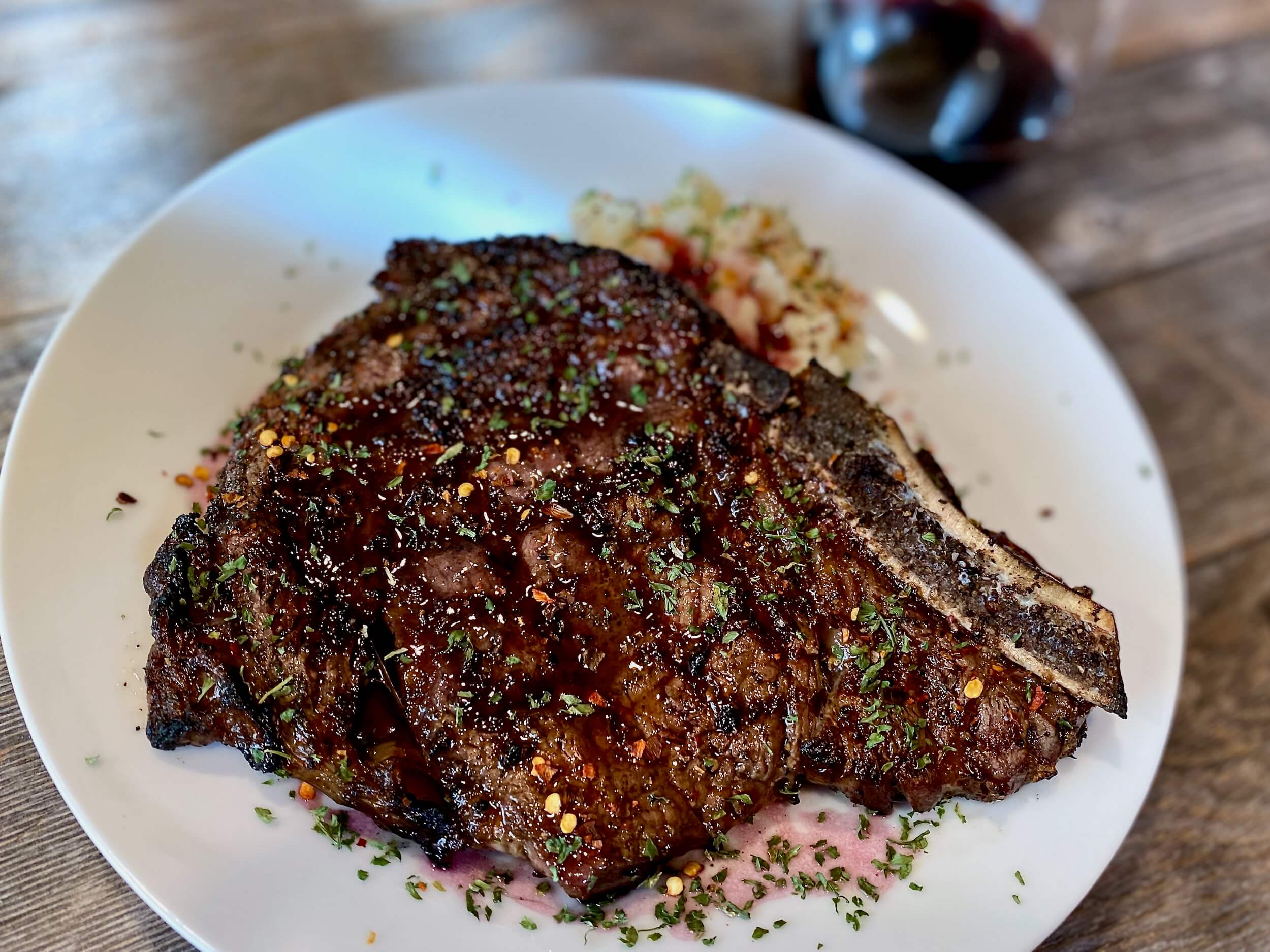 Life At The Table How to Grill Steak. A bone-in grilled ribeye on a white plate garnished with red pepper flakes and parsley with a glass of red wine sitting next to it.