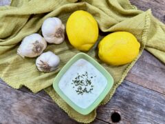Life At The Table Lemon Garlic Aioli. A square bowl of Lemon Garlic Aioli garnished with parsley sitting on a green napkin surrounded with fresh garlic and lemons.