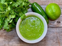 Life At The Table Peruvian Green Sauce. A white bowl filled with Peruvian green sauce surrounded by fresh cilantro, a jalapeno and a lime.