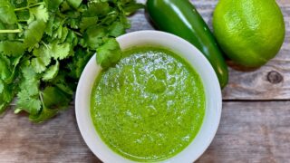 Life At The Table Peruvian Green Sauce. A white bowl filled with Peruvian green sauce surrounded by fresh cilantro, a jalapeno and a lime.