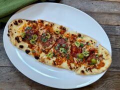 Life At The Table Grilled Flatbread. A grilled flatbread cut into pieces on a white plate sitting on a wooden table with a green napkin beside it.