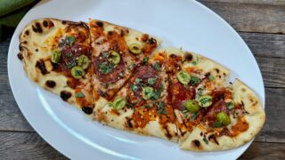 Life At The Table Grilled Flatbread. A grilled flatbread cut into pieces on a white plate sitting on a wooden table with a green napkin beside it.