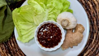 Life At The Table Homemade Hoisin Sauce. A bowl of homemade hoisin sauce on a white plate with butter lettuce, a head of garlic, and some fresh ginger sitting on a wicker charger with a green napkin beside it.