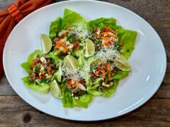 Life At The Table Lettuce Wraps. 5 butter leaves on a white platter filled with cooked ground chicken and garnished with shredded carrots, chopped peanuts, cilantro, and fried rice sticks. There is an orange napkin sitting next to the platter on a wooden table.