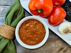 Life At The Table Romesco Sauce. A white bowl of romesco sauce sitting on a wooden table surrounded by a green napkin, red bell pepper, two Roma tomatoes, garlic, and an ancho chile.