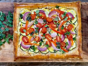 Vegetable Tart Life At The Table. A beautiful, rectangle-shaped vegetable tart with ricotta and parmesan, yellow squash, zucchini, grape tomatoes, red onion, and fresh oregano sitting on a wooden platter with several sprigs of fresh oregano sitting next to it.