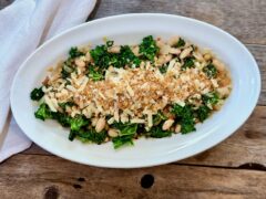 Life At The Table Beans and Greens. An oblong bowl filled with beans and greens garnished with toasted bread crumbs and parmesan on a wooden table with a white napkin beside it.