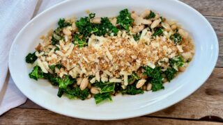 Life At The Table Beans and Greens. An oblong bowl filled with beans and greens garnished with toasted bread crumbs and parmesan on a wooden table with a white napkin beside it.
