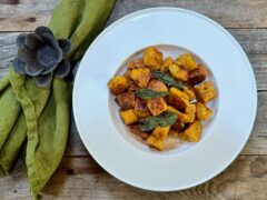 Life At The Table Pumpkin Gnocchi with Brown Butter, Sage, and Garlic Sauce. A white bowl of pumpkin gnocchi garnished with fried sage and edible flowers with a green napkin sitting beside it on a wooden table.