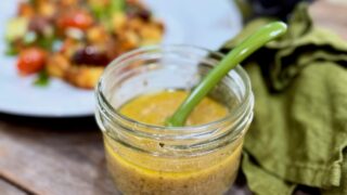 Greek Salad Dressing | Life At The Table. A jar of Greek Salad dressing with a green spoon on a wooden table with a green napkin beside it and a salad behind it.