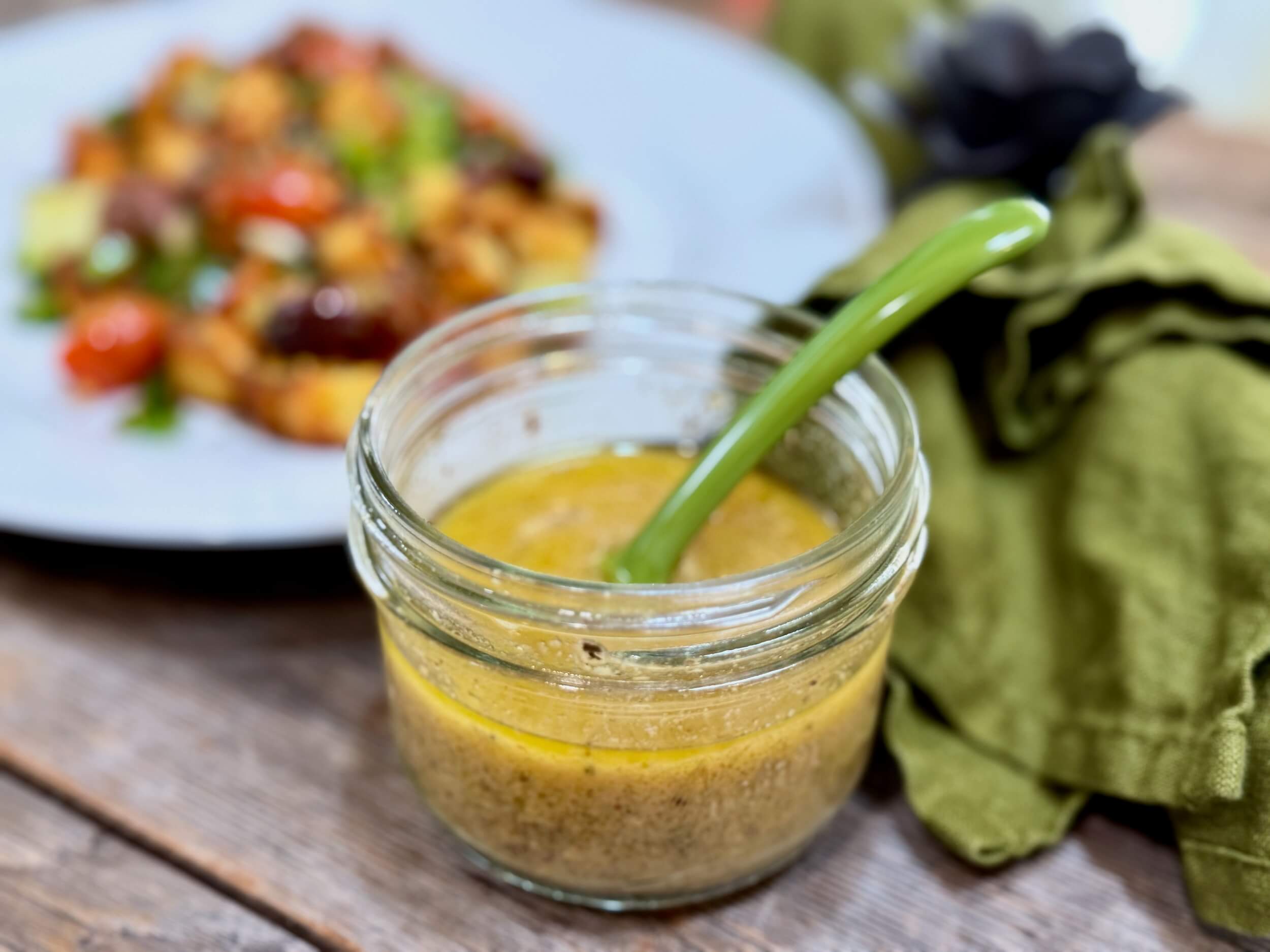 Greek Salad Dressing | Life At The Table. A jar of Greek Salad dressing with a green spoon on a wooden table with a green napkin beside it and a salad behind it.