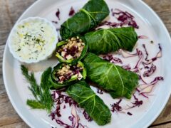Life At The Table Collard Greens Wrap. Five collard greens wraps on a white plate garnished with red cabbage and with a small bowl of a lemon, yogurt, and dill dipping sauce on the side.