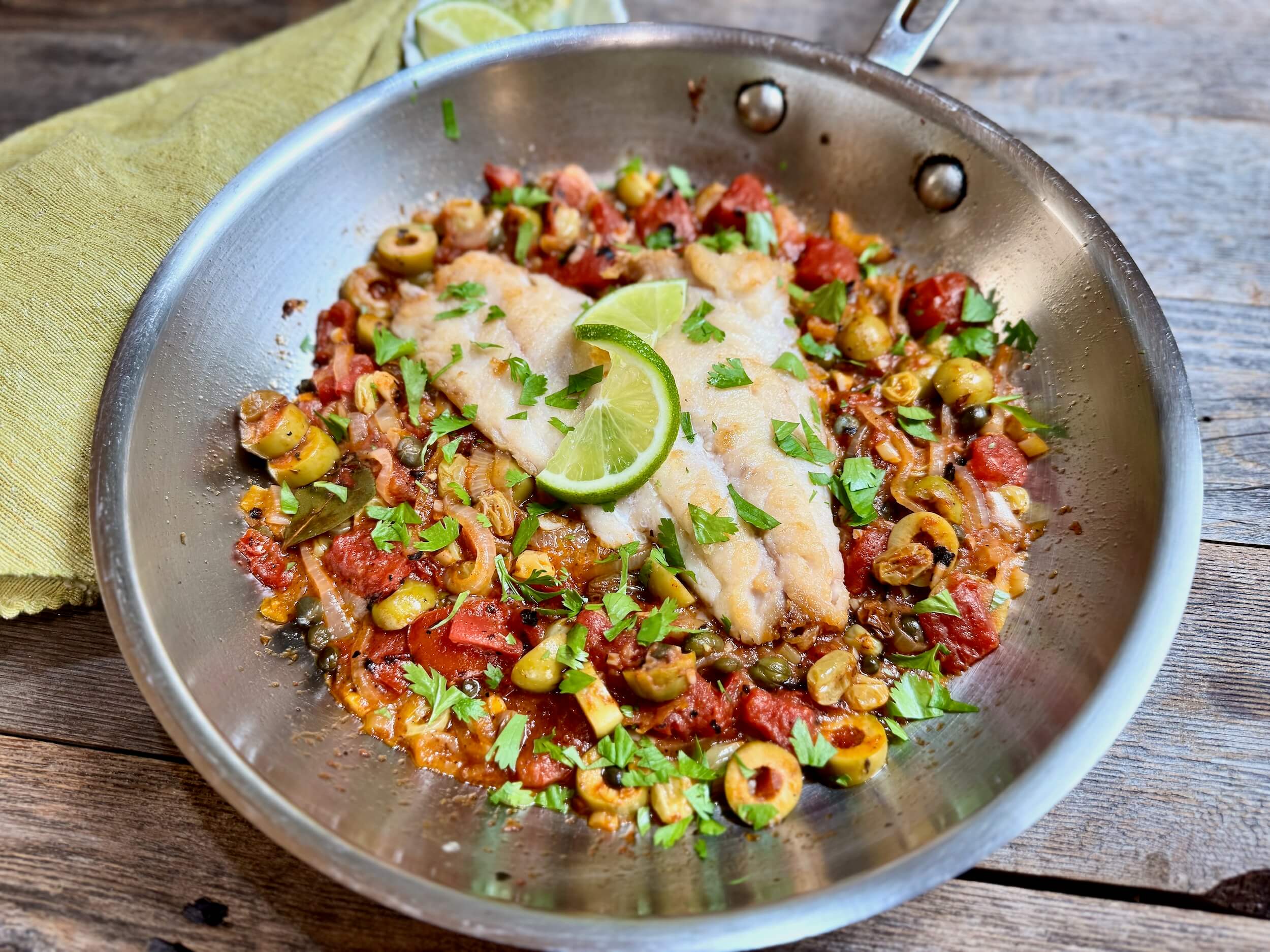 Life At The Table Red Snapper Veracruz. A filet of red snapper nestled in a veracruz-style sauce in a stainless steel skillet with a green napkin and some limes beside it.