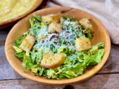 Life At The Table Caesar Salad. A caesar salad in a wooden bowl garnished with croutons, parmesan cheese, and red pepper flakes with extra dressing and a napkin sitting beside it on a wooden table.