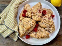 Life At The Table Raspberry Lemon Scones. Raspberry lemon scones stacked on a white plate with fresh raspberries with a checked-napkin and a fresh, whole lemon sitting next to it.