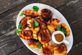 Life At The Table Grilled Peaches. A platter of grilled peaches garnished with mint leaves with a small container of bourbon sauce next to it sitting on a wooden table.