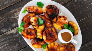 Life At The Table Grilled Peaches. A platter of grilled peaches garnished with mint leaves with a small container of bourbon sauce next to it sitting on a wooden table.