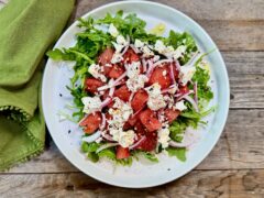 Life At The Table Watermelon and Feta Salad. A watermelon and feta salad with red onion, sumac, and a red pepper garnish on a white platter with a green napkin beside it on a wooden table.