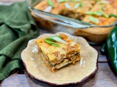Life At The Table Chiles Rellenos Casserole. A slice of chiles rellenos casserole on a small plate sitting in front of a baking dish with fresh poblanos and a green napkin on either side.
